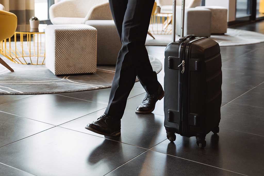 Ein Mann im dunklen Anzug läuft durch das Foyer eines Hotels und zieht einen dunklen Trolley neben sich her.