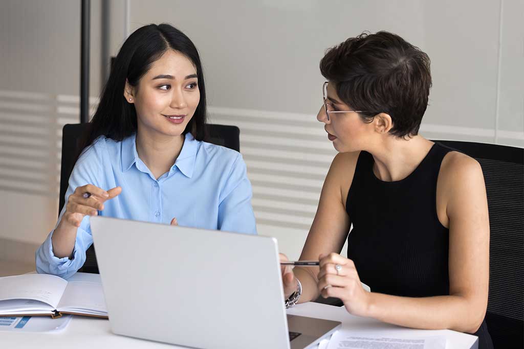 Zwei junge Frauen unterhalten sich vor einem geöffnetem, grauen Laptop.