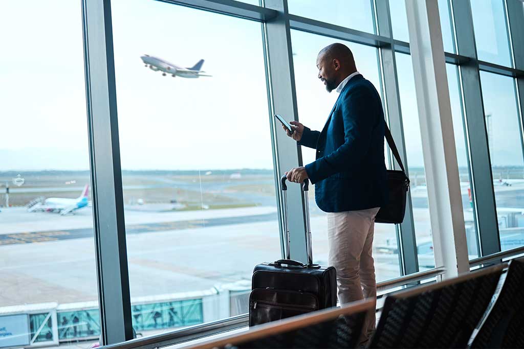 Ein Mann in einem dunkelblauen Anzug steht an einem Flughafen-Fenster. Im Hintergrund startet ein weißes Flugzeug während er auf sein Smartphone schaut.